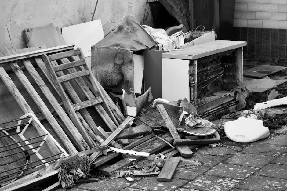 Outdoor scene showing a pile of discarded household items and debris on a paved area adjacent to a building, including wooden furniture panels, a broken wooden ladder, a white plastic object, and various pieces of damaged or discarded material. Behind the debris, there is a partially open storage unit or cabinet with signs of damage, and nearby, a brick wall is visible. The area appears cluttered and unkempt, with some items possibly awaiting disposal or removal. Natural light illuminates the scene, and the setting suggests an outdoor collection point for bulky waste or debris removal, which could be relevant to home relocation or furniture transport services provided by Man with Van Eel Pie Island.