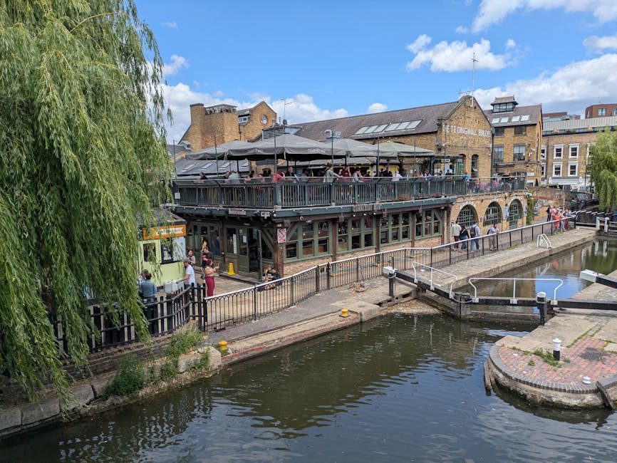 A large multi-level building with an outdoor terrace surrounded by a metal railing, situated alongside a narrow river or canal. The terrace is covered with large grey umbrellas and is busy with people seated, enjoying the view. The building has a brick exterior with arched windows on the ground floor and windows on the upper level, some of which are skylights. To the left, a green vintage van is parked near the pavement where a few individuals are standing or walking, carrying boxes or bags as part of a home relocation or moving process. The scene includes a black safety fence along the water’s edge and a small boat moored close to the quay. The background features multi-storey brick buildings and a partly cloudy sky, with lush green trees adding to the scene. This setting illustrates a typical environment for house removals or furniture transport in a historic riverside area, supported by the presence of the removal service company, Man with Van Eel Pie Island.