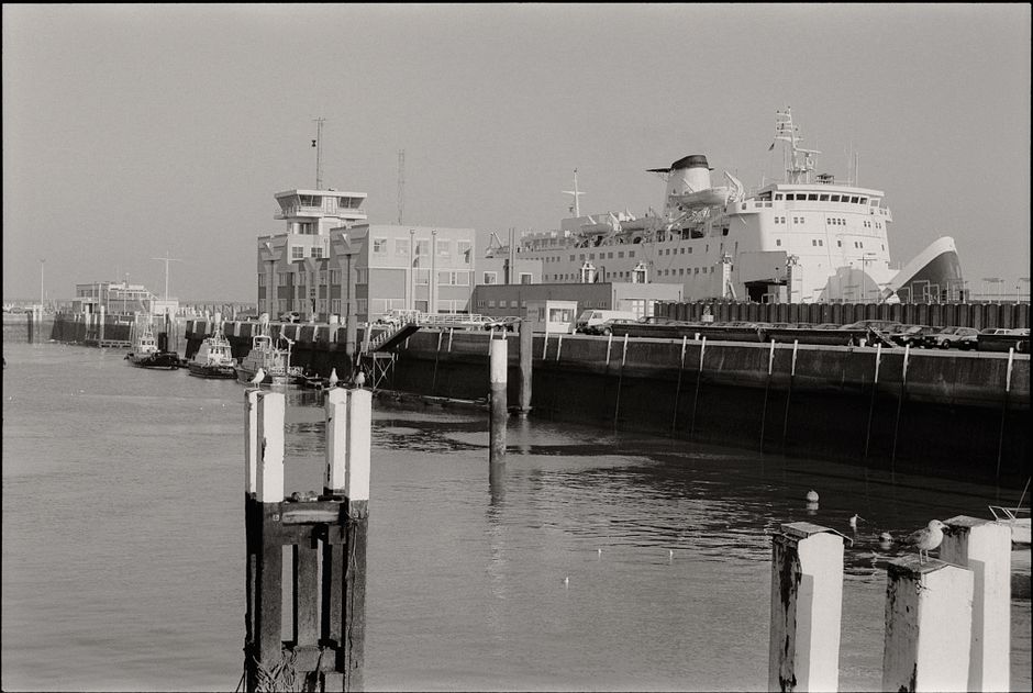 Two ferry boats docked at the quay under a bright blue sky with wispy white clouds. The ferries are painted white and yellow, each featuring a prominent yellow funnel with a blue stripe, and are label