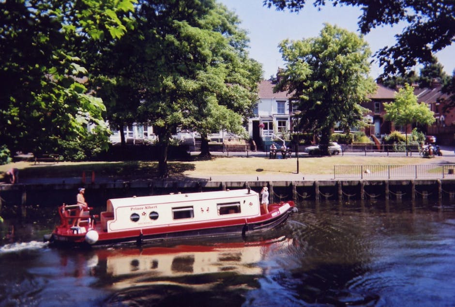 A red and white narrowboat named 'Prince Albert' is navigating along the waterway adjacent to a residential area, with two people on board. The boat is fitted with windows and a small deck at the rear, where a person is standing. The water reflects the boat's colors and nearby trees. On the opposite bank, there is a grassy area with large trees providing shade and several houses with front gardens, some with visible pathways and railings. The scene is outdoors during daylight, with clear weather and bright natural light illuminating the scene. This image depicts aspects of transportation and leisure on the canal, which aligns with house removals and moving services, emphasizing the importance of logistical planning in home relocation on London's Eel Pie Island, as provided by Man with Van Eel Pie Island.