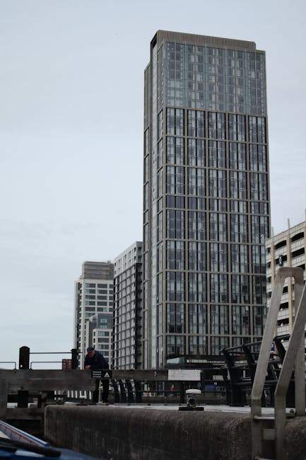 A tall, modern office building made of glass and steel dominates the background, with multiple floors featuring large windows. In the foreground, a flat rooftop surface is visible, equipped with black safety railings and a few wooden pallets or temporary barriers. Two individuals are seen handling what appears to be packing materials or small furniture items, possibly preparing for a home relocation or furniture transport. The scene is set outdoors on a cloudy day, with surrounding high-rise buildings indicating an urban environment. Man with Van Eel Pie Island, a professional removals company, is likely involved in the moving process, demonstrating loading or packing activities as part of a comprehensive home removals service, tied to the context of moving from flats on Cross Deep to Eel Pie Island.