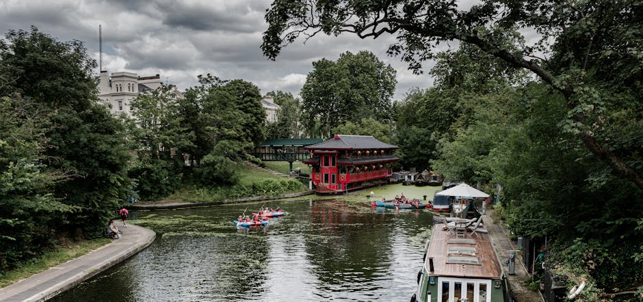 A scenic view of the River Thames near Eel Pie Island, with lush green trees lining the riverbank and a cloudy sky overhead. In the foreground, a narrow paved path runs along the water's edge, where a person is walking and another is sitting on a bench. Several small boats with red, blue, and white pontoons are visible on the river, some being paddled by individuals engaged in a leisure activity. On the right side, a narrowboat with a weathered roof is moored along the bank, and nearby, a small outdoor seating area with a white canopy tent is set up, possibly for a local event or resting spot for visitors. In the background, a distinctive red and black traditional Asian-style building with a decorative rooftop extends over the water, supported by stilts, and behind it, a dense canopy of mature trees provides shade. The overall scene captures the tranquil environment of this riverside location, which is relevant to house removal or relocation services on Eel Pie Island, as provided by Man with Van Eel Pie Island.